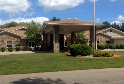 Exterior view of a single-story brick building with a covered entrance supported by stone pillars, surrounded by green grass, shrubs, and trees under a partly cloudy blue sky.