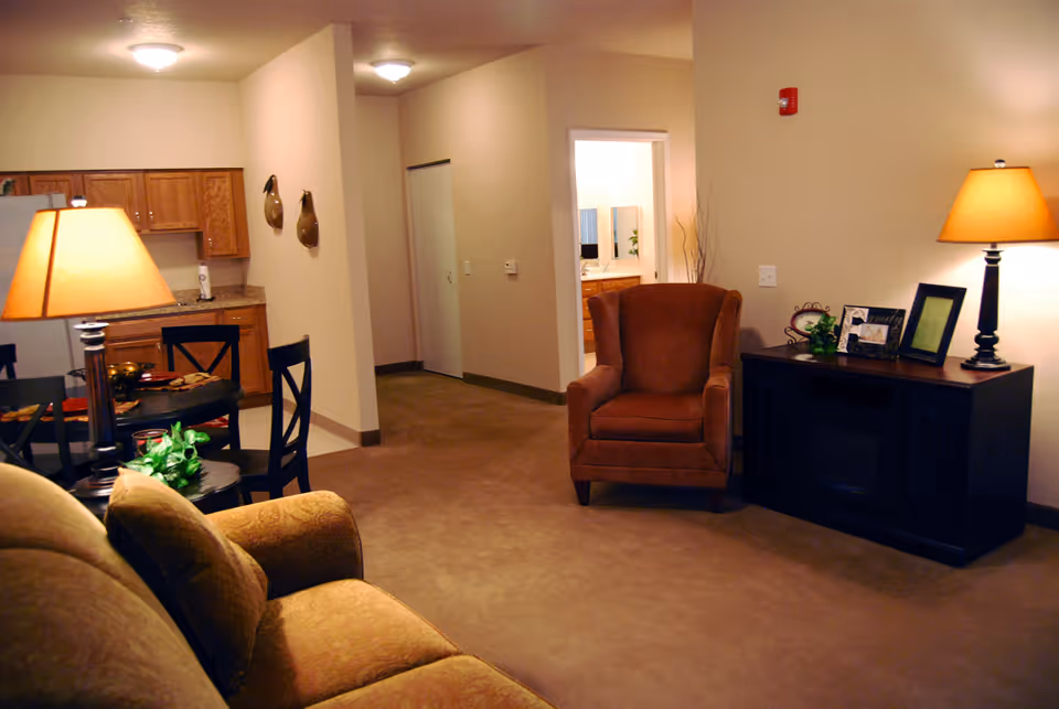 Interior view of a senior living facility showing a cozy living room area with a brown armchair, a dark wooden cabinet with a lamp and picture frames on top, and a beige couch partially visible. In the background, there is a dining area with a round table and chairs, a kitchen with wooden cabinets, and a hallway leading to another room with a sink and mirror visible.