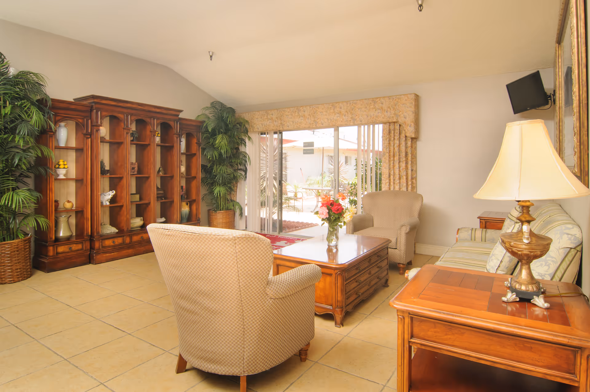 A cozy living room with beige tiled floors, a wooden coffee table with a vase of flowers, two upholstered armchairs, a striped sofa, a wooden side table with a lamp, a large wooden bookshelf with decorative items, and large sliding glass doors leading to an outdoor patio area.