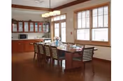 A dining room with a long wooden table surrounded by eight chairs. The table is set with plates, glasses, and a floral centerpiece. The room has wooden flooring, large windows with blinds, and wooden cabinets with glass doors along one wall. A modern chandelier hangs above the table.