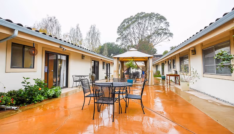 Courtyard with metal patio tables and chairs and a central gazebo between single-story buildings on a wet paved surface.