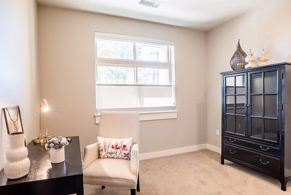 Bright small sitting room with an upholstered armchair and floral pillow beside a desk and a dark glass-front cabinet under a window.