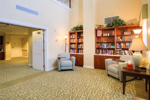 A cozy interior common area with two light blue armchairs, a wooden side table with a lamp and tissue box, and two wooden bookshelves filled with books and decorative items. The room has beige walls, a patterned carpet, and an open doorway leading to a hallway.