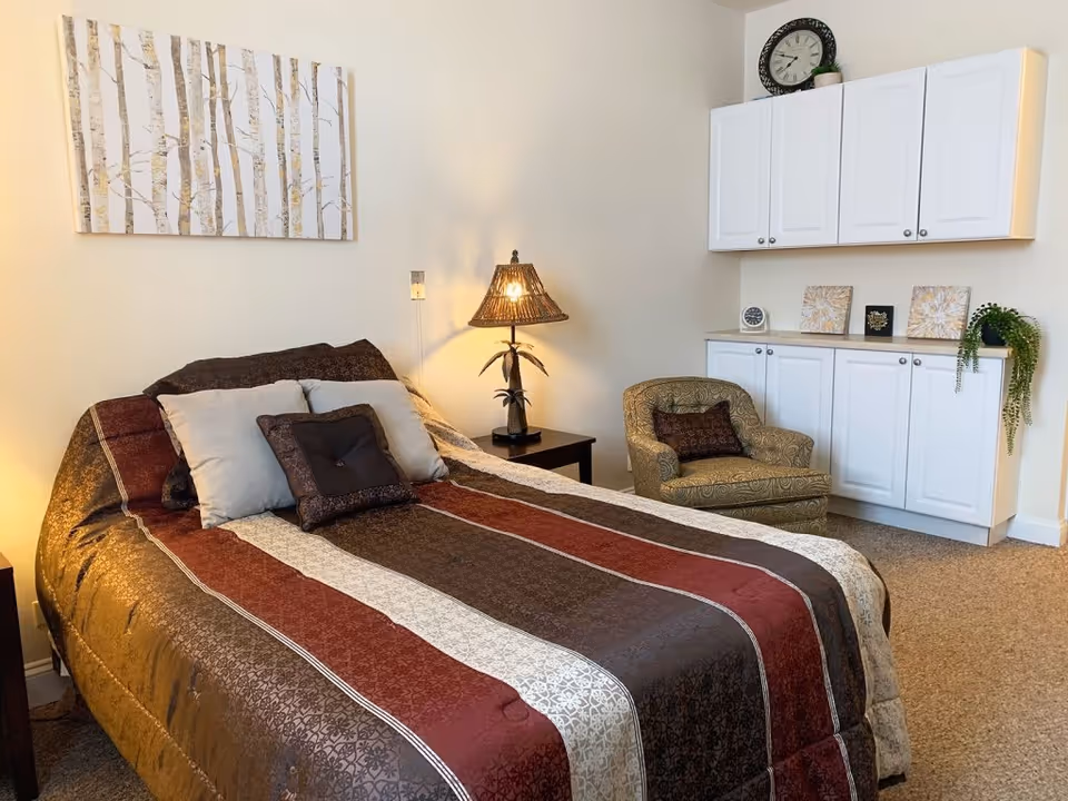 A cozy bedroom with a bed covered in a striped brown, red, and beige comforter with three pillows. To the right of the bed is a small table with a decorative lamp. On the far wall, there is a white cabinet with a clock and decorative items on top, and a patterned armchair with a small pillow. A painting of birch trees hangs on the wall above the bed.