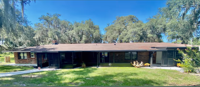 Single-story brick building with a brown roof surrounded by green grass and large trees under a clear blue sky.