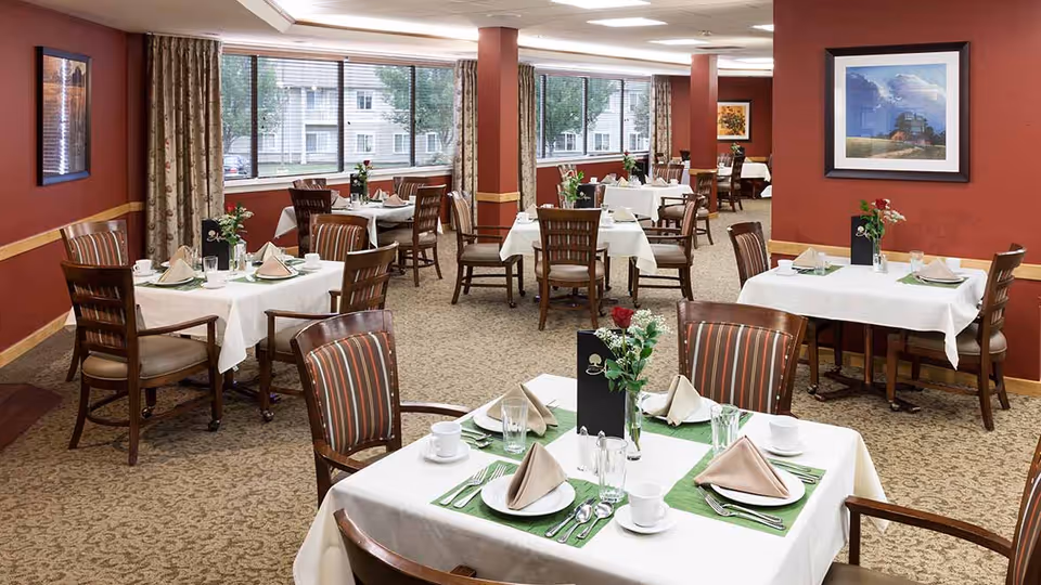A dining room with multiple tables covered in white tablecloths, each set with plates, cups, silverware, and folded napkins. The chairs have wooden frames with striped cushions. The room has red walls with framed artwork and large windows letting in natural light.