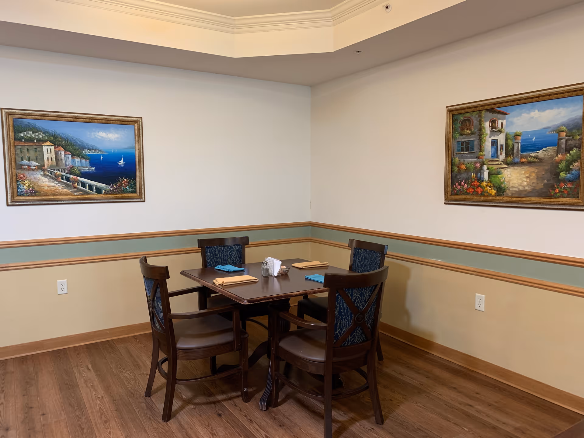 A small dining area with a square wooden table set for four, surrounded by four wooden chairs with blue patterned cushions. The room has light-colored walls with a green and wood trim, wooden flooring, and two framed paintings depicting coastal scenes hanging on the walls.