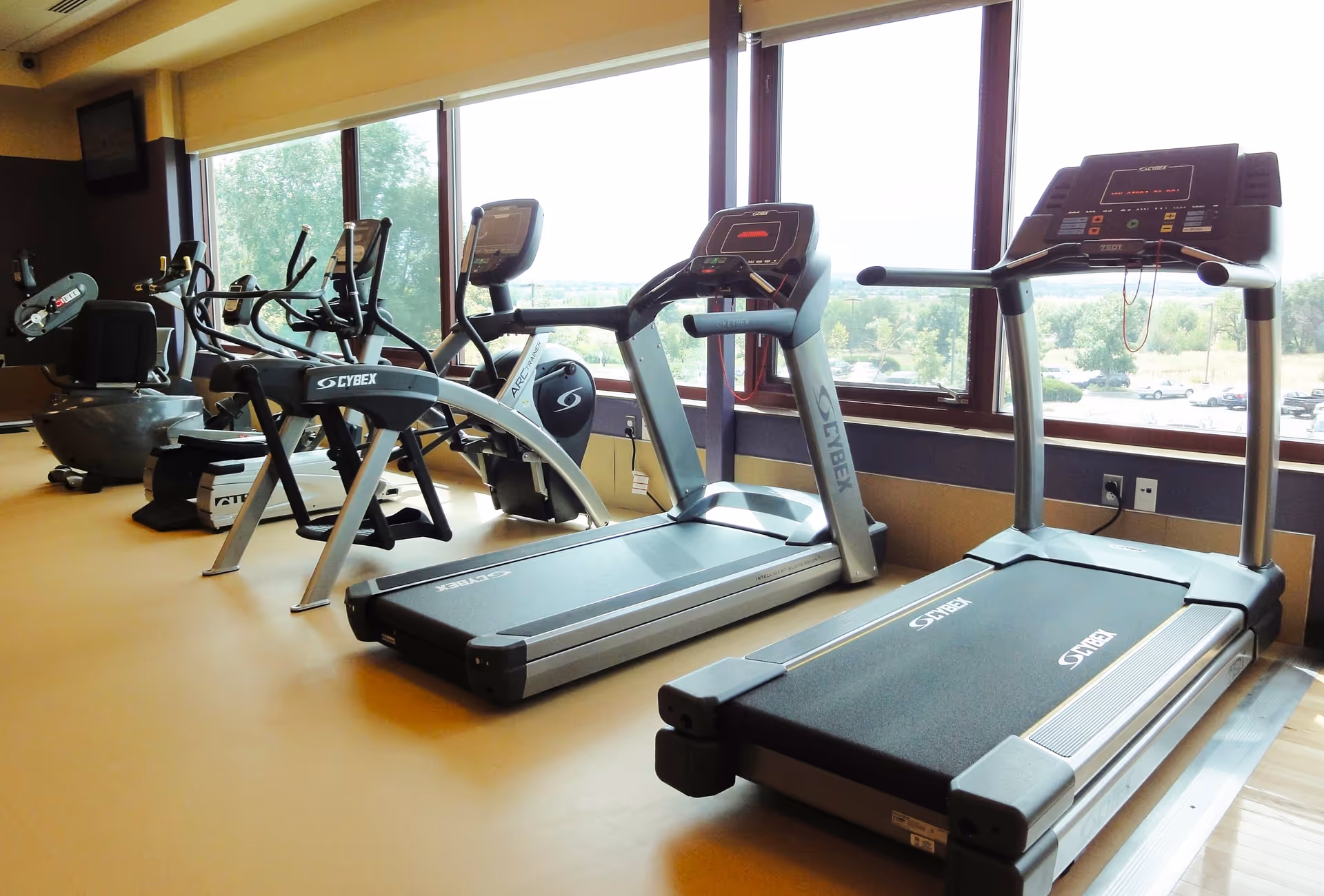 A fitness room with several exercise machines including treadmills, elliptical trainers, and stationary bikes positioned in front of large windows overlooking a green outdoor area with trees and parked cars.