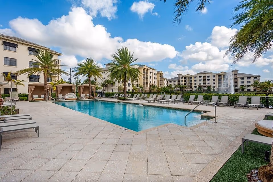 Outdoor swimming pool area with lounge chairs, cabanas, palm trees, and multi-story residential buildings under a blue sky.