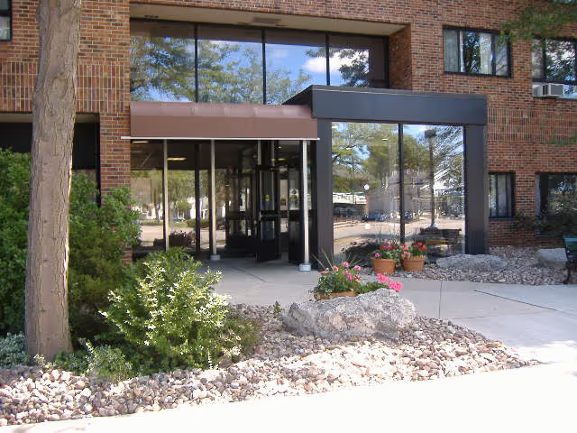 Entrance to a brick building with large glass windows and a glass door. There are plants, rocks, and a tree in front of the entrance, with a concrete walkway leading up to the door.
