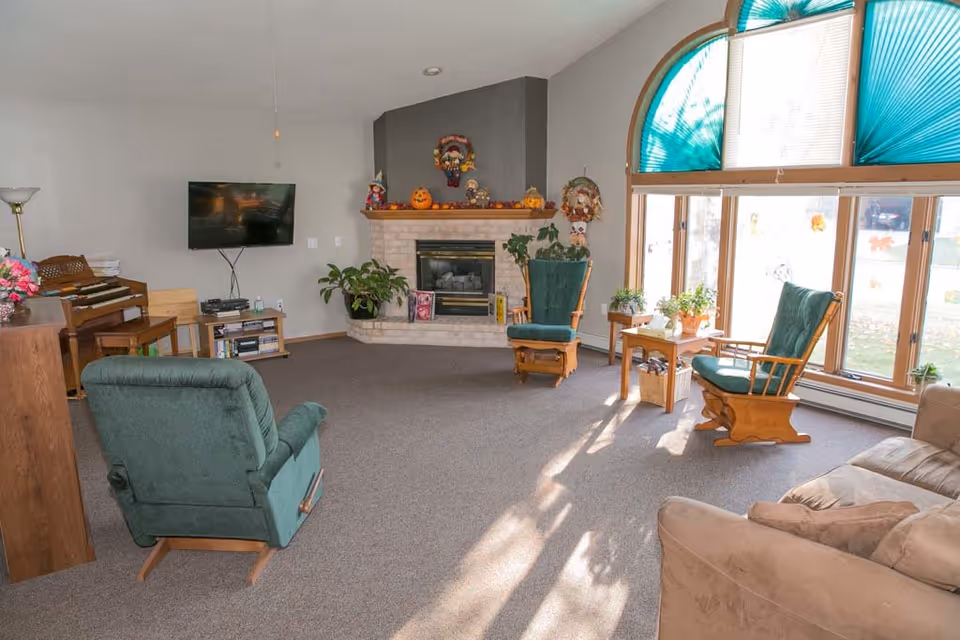 A cozy living room with a beige couch, two green cushioned wooden rocking chairs, a green recliner, a brick fireplace decorated with autumn-themed items, a wall-mounted TV, a piano, and large windows with teal blinds letting in sunlight.
