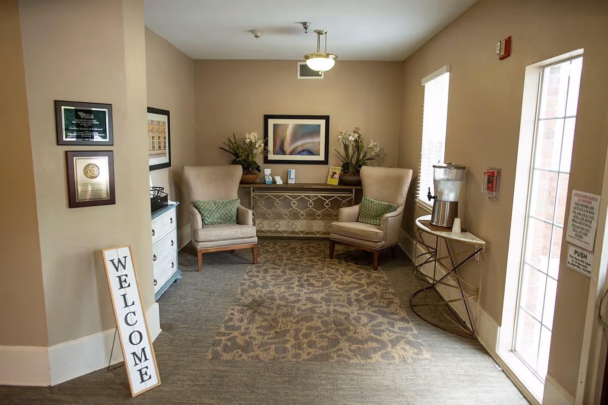A cozy interior seating area with two beige armchairs with green patterned pillows, a decorative console table with plants and framed artwork above it, a patterned area rug on the floor, a water dispenser on a small round table near a glass door, and a vertical 'WELCOME' sign leaning against a wall.