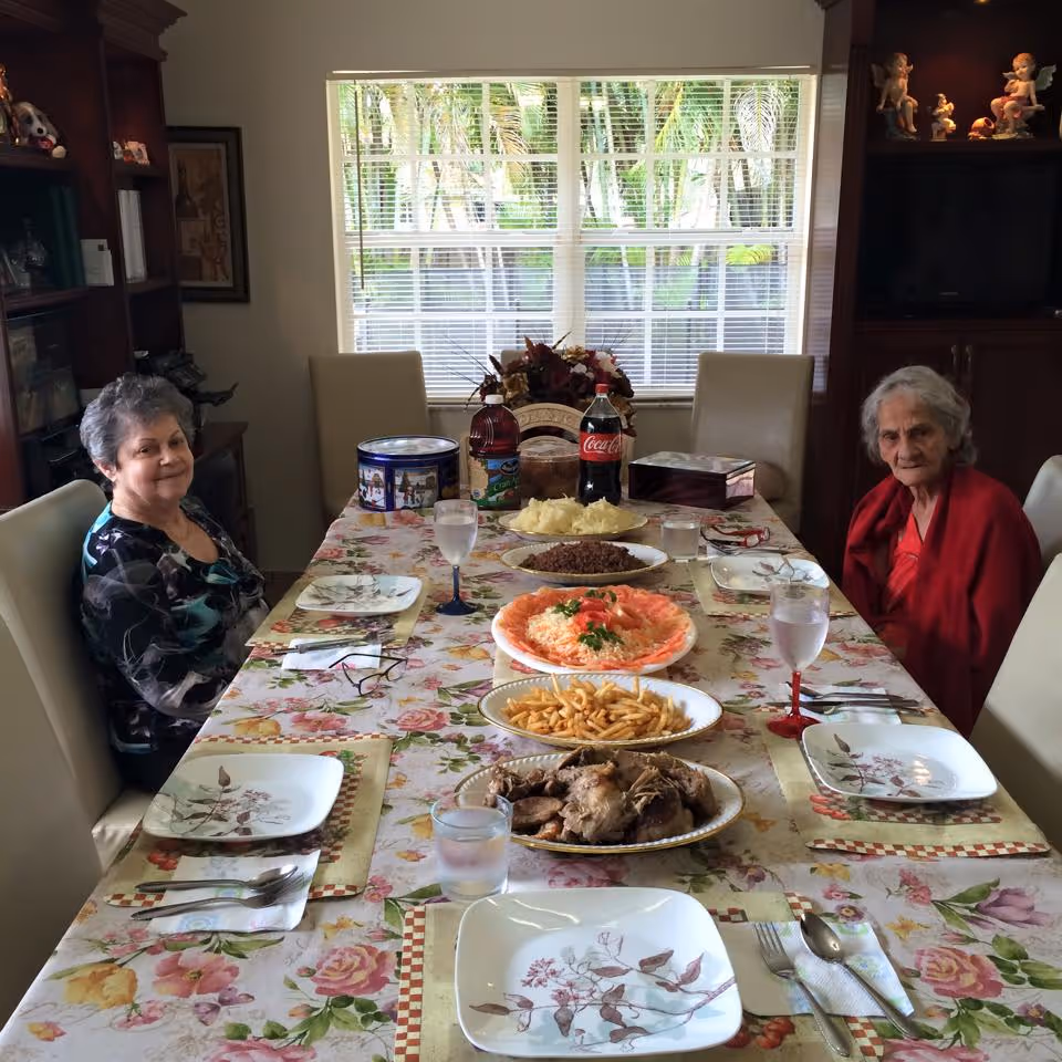 Two elderly women sitting at a dining table set with plates, glasses of water, and various dishes including mashed potatoes, ground meat, fries, a salad, and roasted meat. The table is covered with a floral tablecloth and is in a room with a large window showing greenery outside. There are shelves with decorative items and a TV in the background.