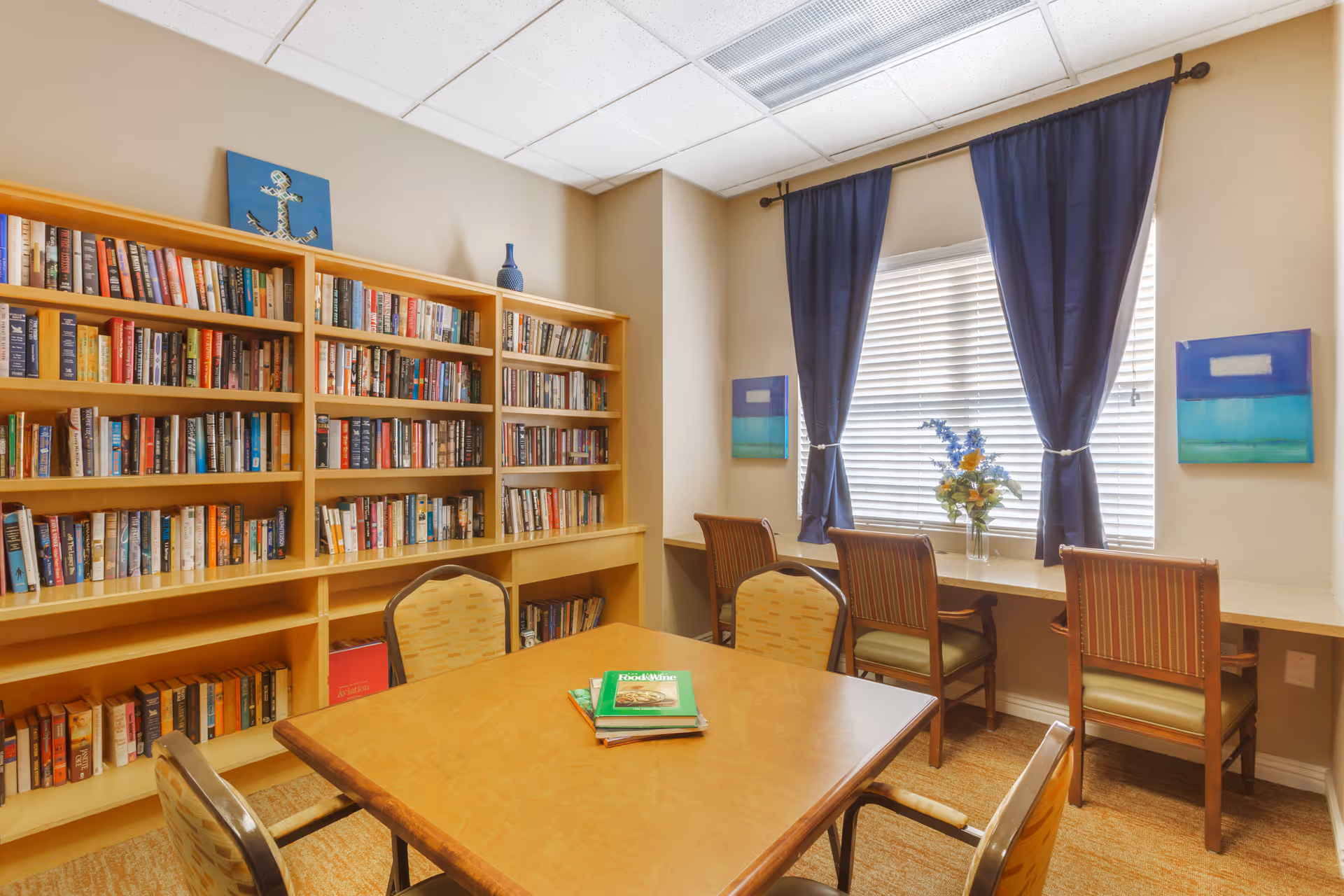 A cozy reading room with a wooden table and four chairs in the center. Behind the table is a large bookshelf filled with books. To the right, there is a window with blue curtains and a windowsill with three chairs and a vase of flowers. Two abstract paintings hang on the wall beside the window.