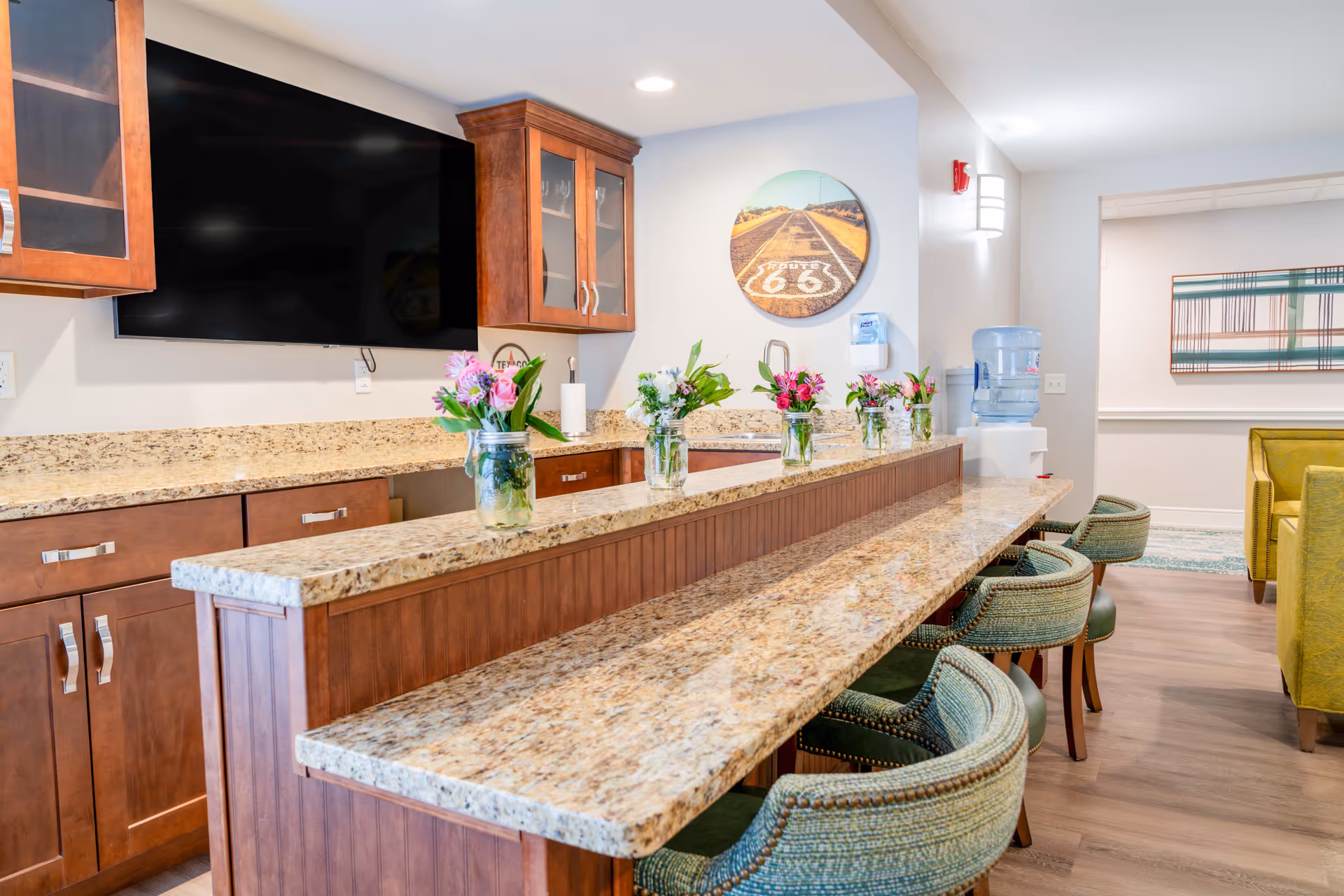 Well-lit communal kitchen/bar area with granite countertops, cabinets, barstools, and vases of flowers.