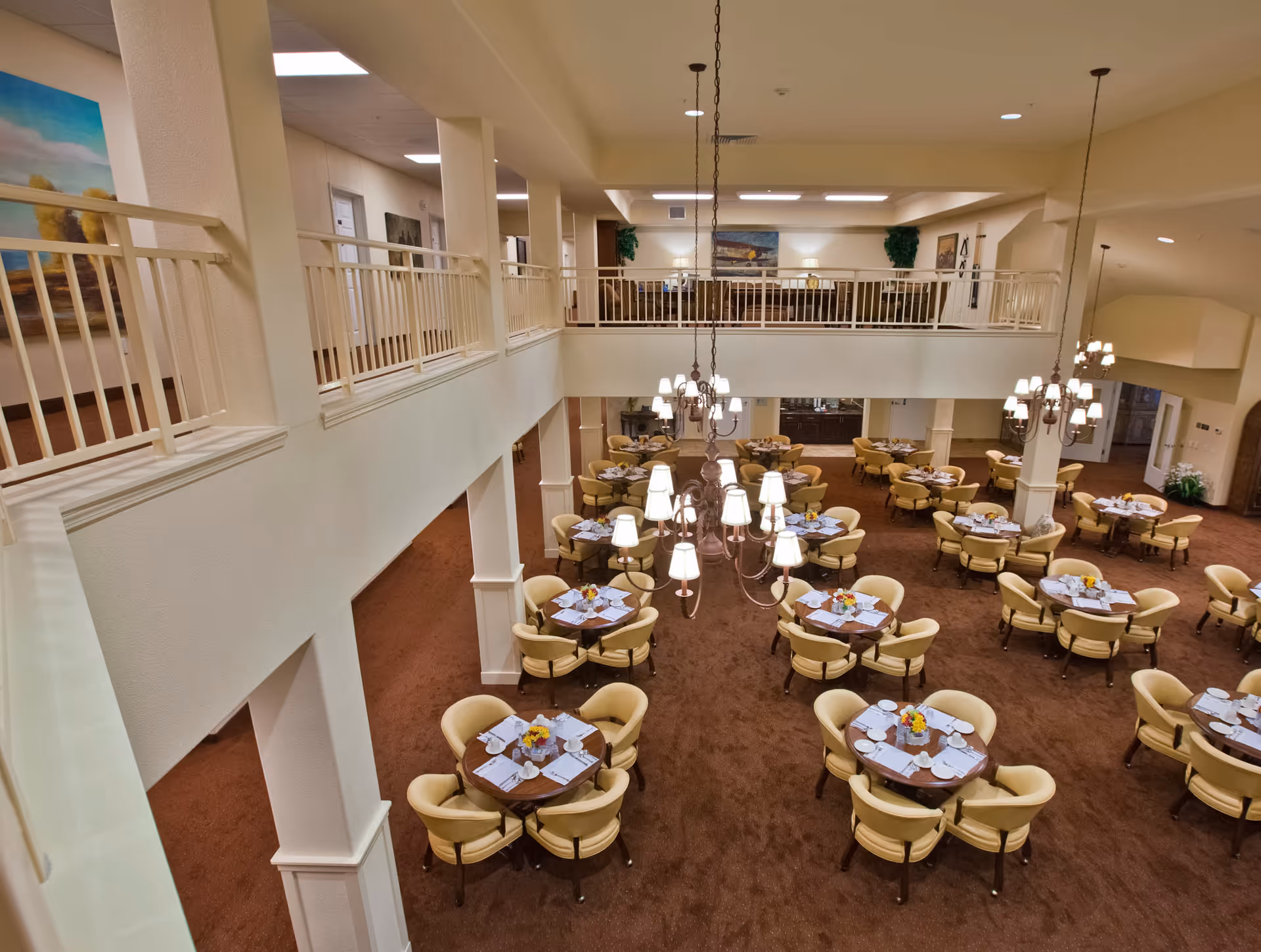 Large two-story dining room with round tables set for meals, upholstered chairs, and hanging chandeliers.