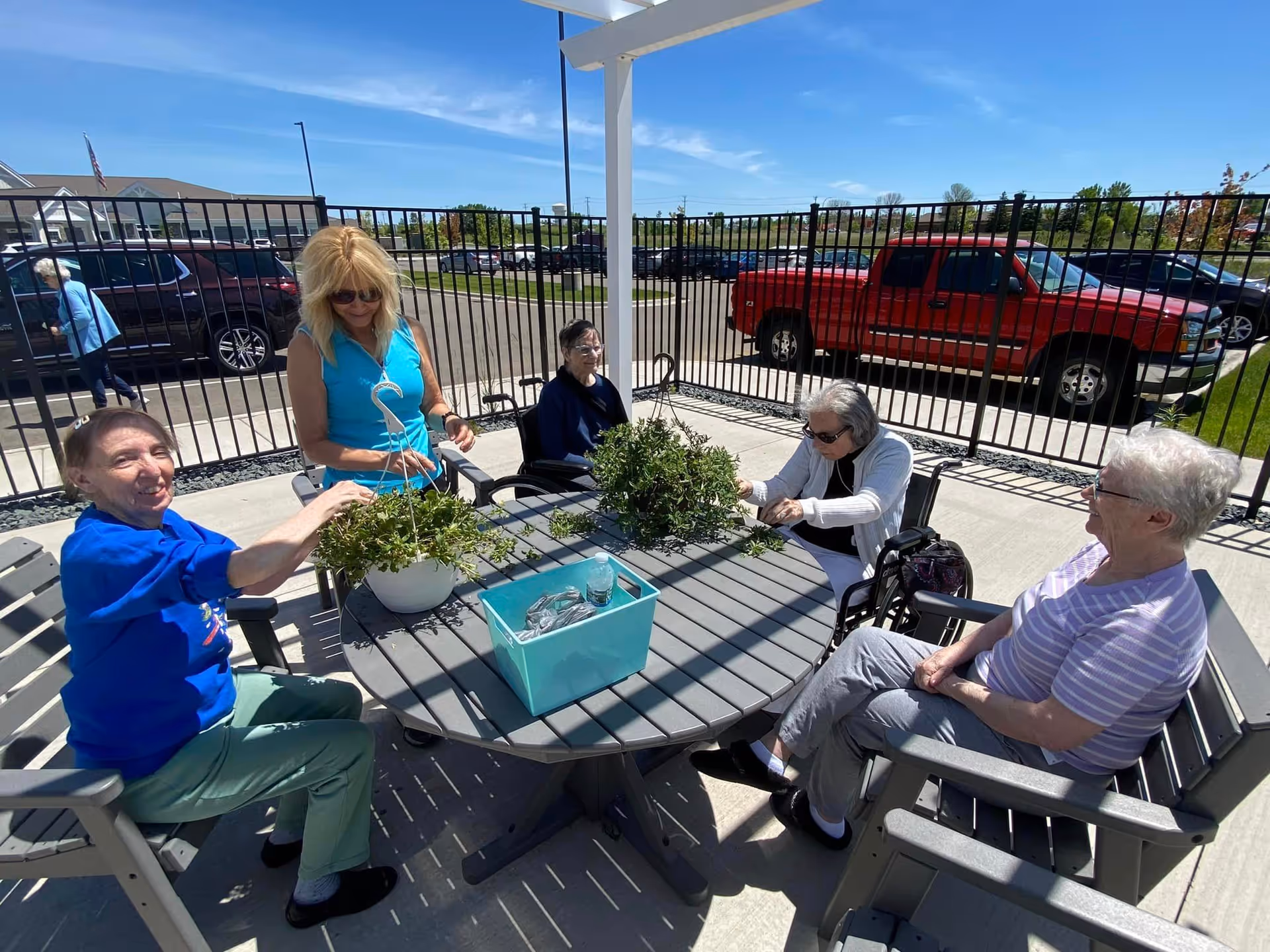 Five elderly women sitting around a round outdoor table under a pergola, tending to potted plants. They are in a fenced patio area with parked cars visible in the background under a clear blue sky.