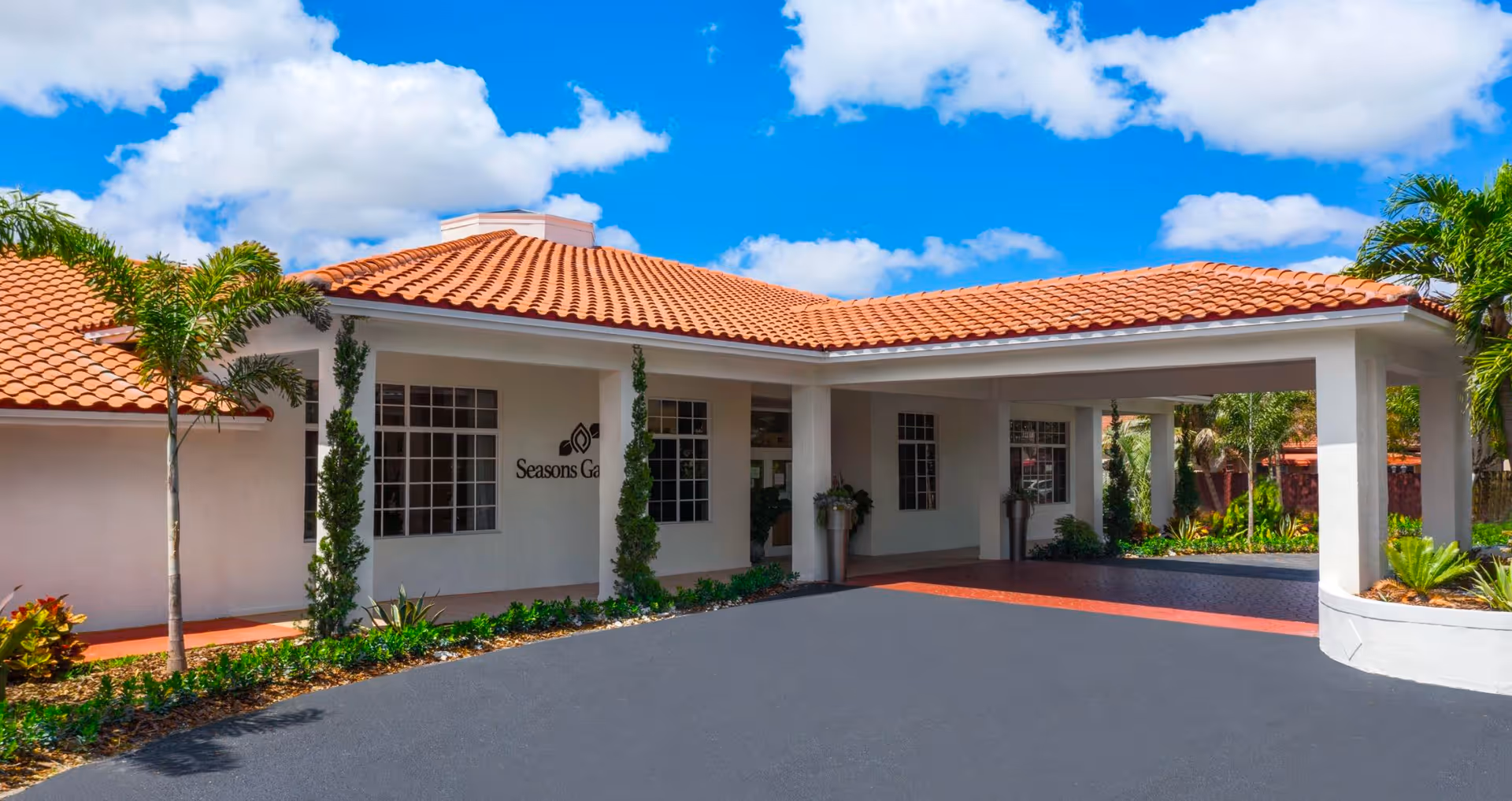 Exterior view of a single-story building with a red tile roof and white walls under a bright blue sky with scattered clouds. The building has large windows and a covered entrance area with columns. Palm trees and landscaped plants surround the entrance. The name 'Seasons Gardens' is visible on the wall near the entrance.