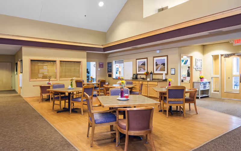 A bright and spacious common area in an assisted living facility with several wooden tables and chairs arranged for dining or socializing. Each table has a small vase with flowers. The room has high ceilings, beige walls, and large windows allowing natural light. There are framed pictures on the walls and a sideboard with decorative items. Doors and hallways lead to other parts of the facility.