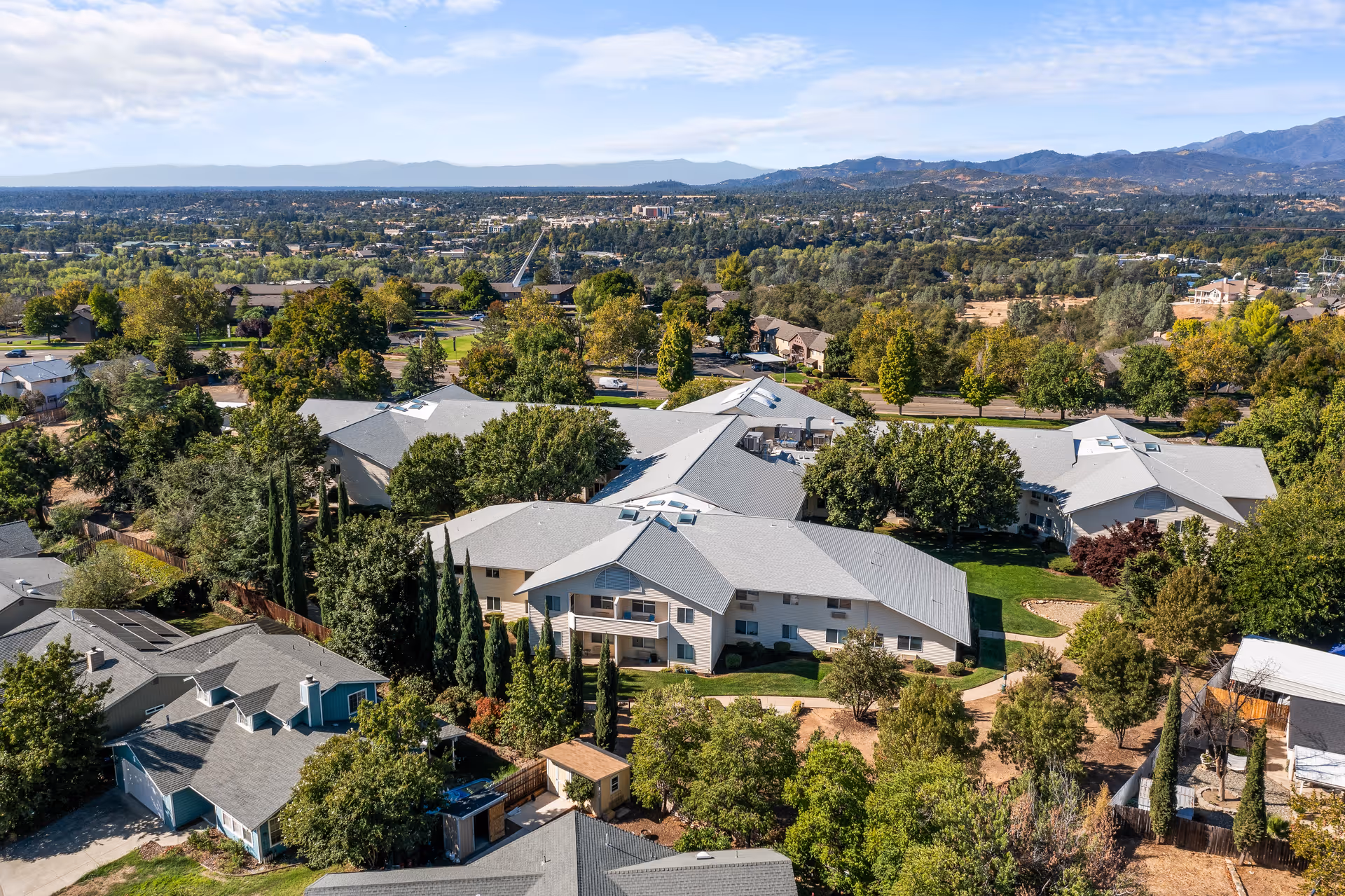 Aerial view of Hilltop Estates - A Provincial Senior Living Community showing multiple buildings with gray roofs surrounded by trees and greenery, with a backdrop of distant mountains and a clear sky.