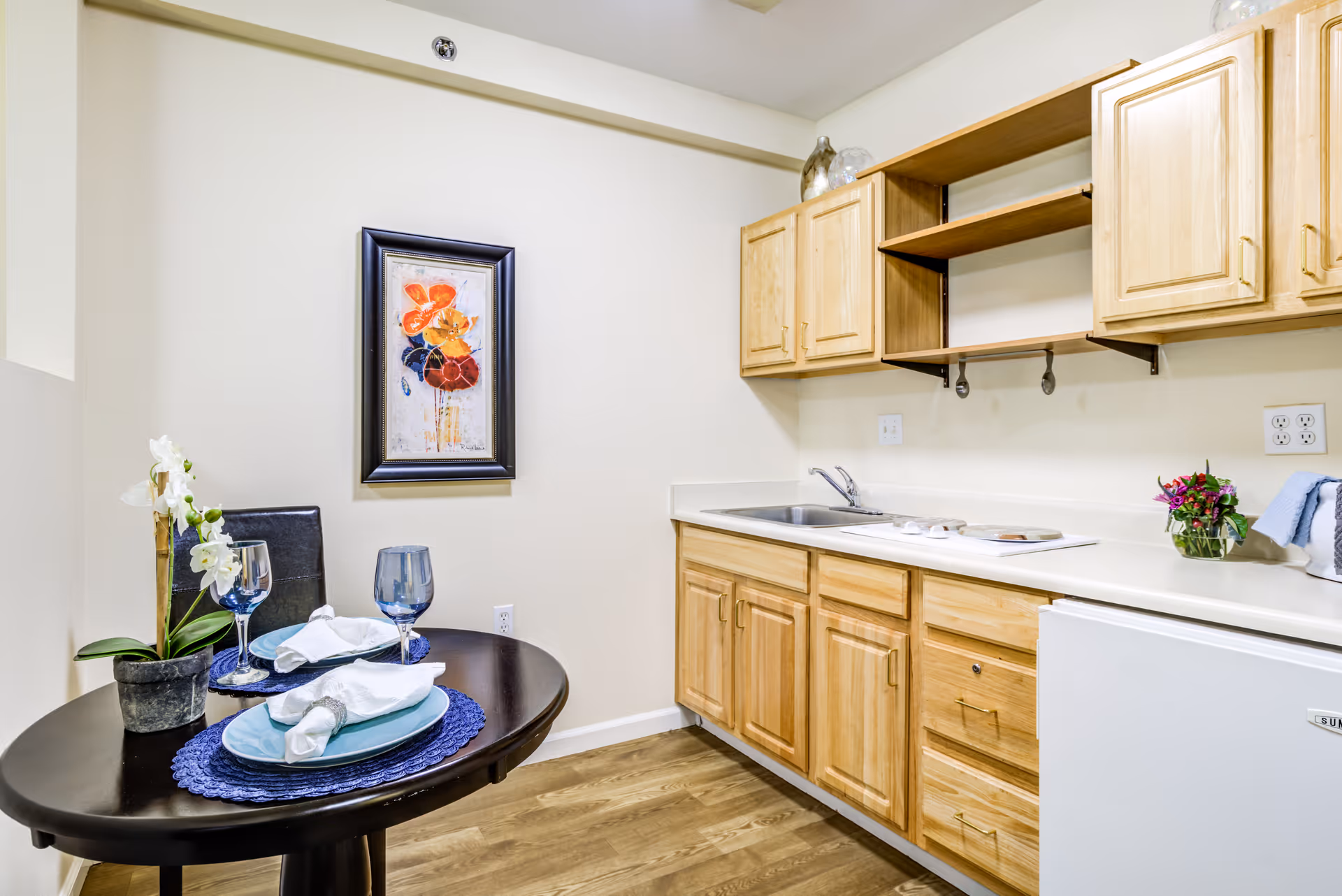 Small kitchen area with light wood cabinets, a white countertop, a sink, and a mini refrigerator. A round dark wood table is set for two with blue placemats, plates, napkins, and glasses. A potted orchid and a framed floral painting are on the wall.