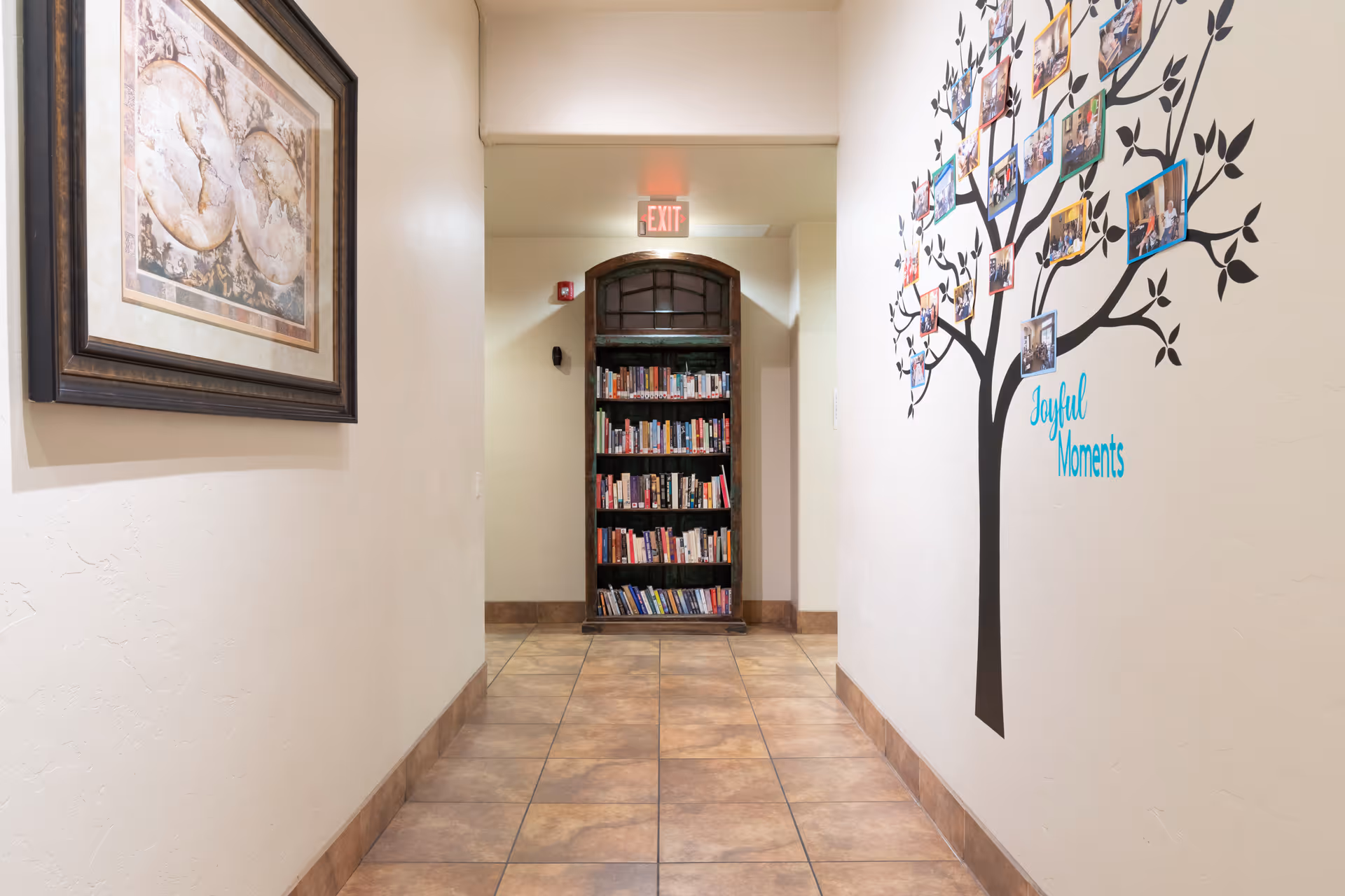 A hallway with tiled floor and cream-colored walls. On the left wall, there is a framed artwork. On the right wall, there is a decorative tree decal with photos attached and the words 'Joyful Moments'. At the end of the hallway, there is a bookshelf filled with books beneath an arched window, and an illuminated exit sign above.