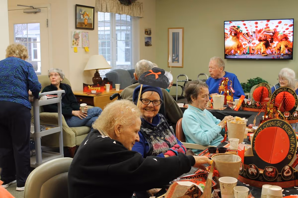 A group of elderly people sitting around tables in a common room decorated for Chinese New Year, with festive red and gold decorations and food containers. Some are engaged in conversation, and a television screen in the background displays a colorful image. The room has a cozy atmosphere with chairs, a lamp, and wall art.
