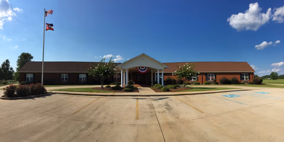 Front exterior view of Atwood Personal Care Home, a single-story brick building with a covered entrance supported by white columns. Two flagpoles with American and Texas flags stand on the left side, and there are landscaped bushes and trees around the entrance. The sky is clear with a few clouds, and there are marked handicap parking spaces in the concrete driveway.