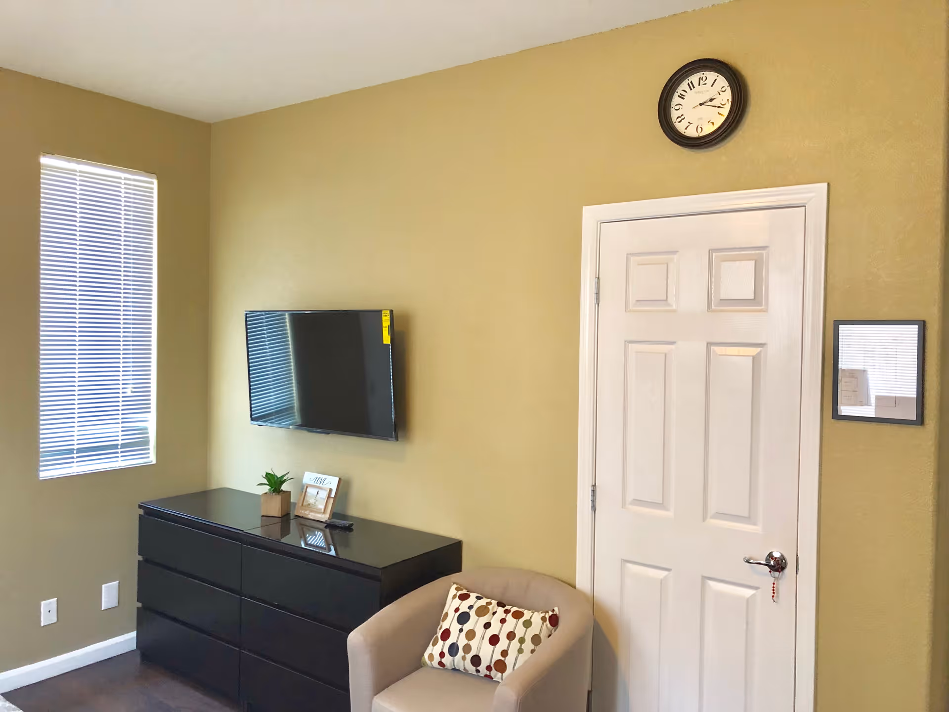 A small room with beige walls featuring a wall-mounted flat-screen TV above a black dresser. To the right, there is a white door with a round clock above it and a framed item on the wall. A beige armchair with a decorative pillow is placed in front of the door. A window with closed blinds is on the left wall.