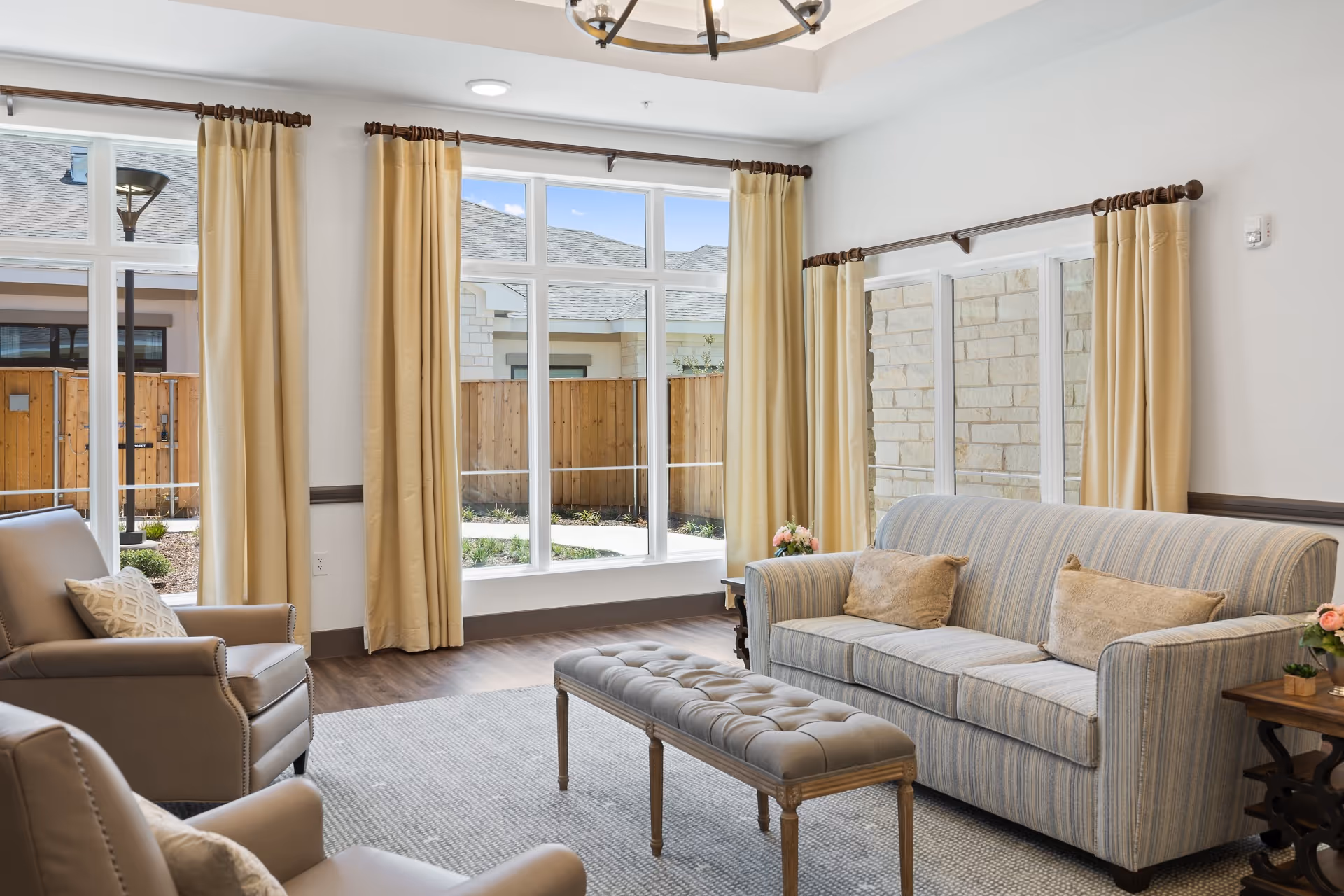 A bright and cozy living room with large windows covered by beige curtains, a striped sofa with two beige pillows, two beige armchairs with patterned pillows, a tufted bench, and wooden side tables with small flower arrangements. The room has wooden flooring and a light gray area rug.