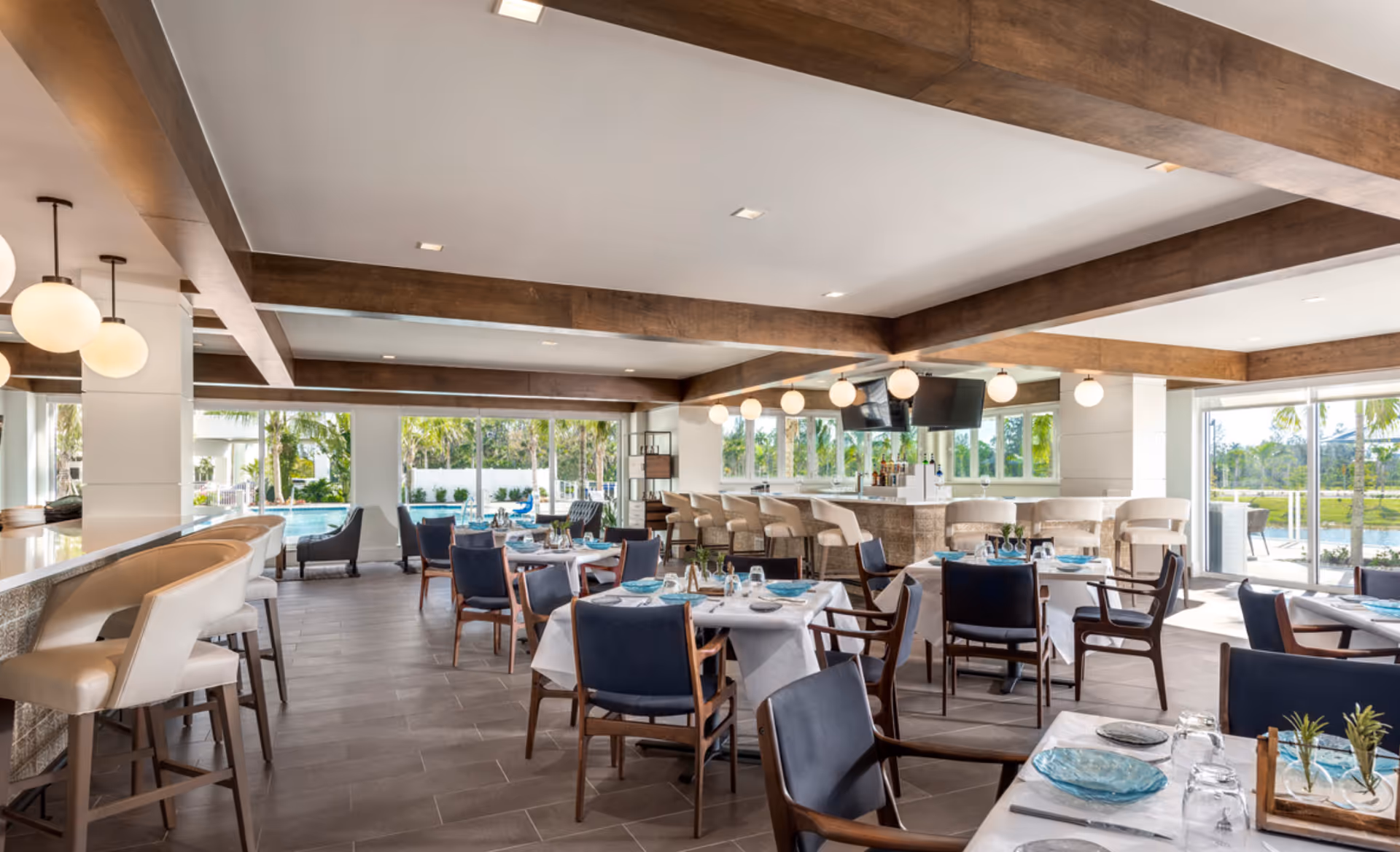 A bright and modern dining area with tables set with blue glass plates, glasses, and silverware. The room features wooden ceiling beams, large windows with views of palm trees and a pool outside, and a bar area with white cushioned bar stools and hanging globe lights.