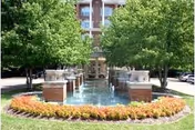 A landscaped outdoor area with a rectangular water feature surrounded by brick pillars and trees, with a multi-story building visible in the background.