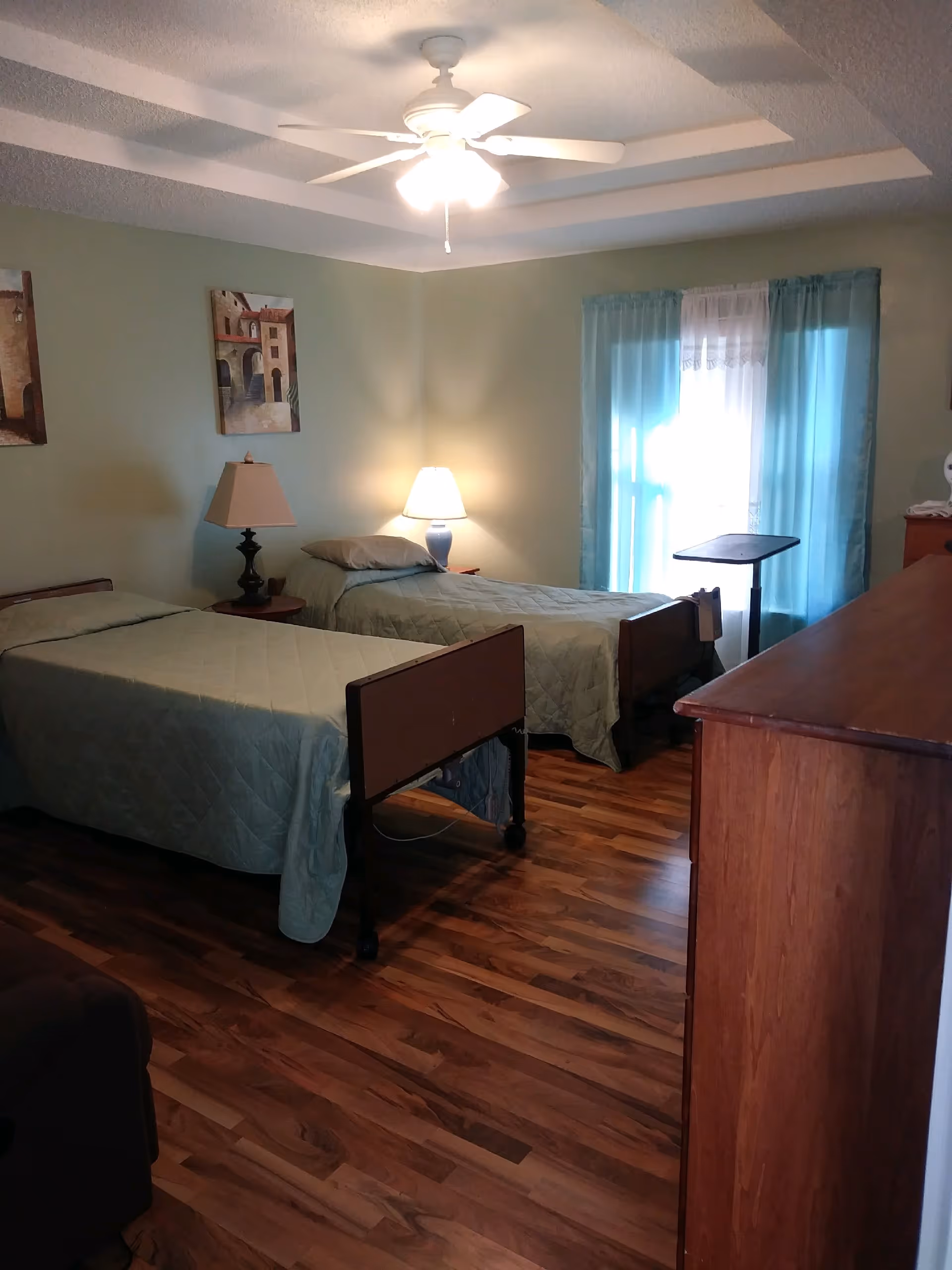 A bedroom with two single beds covered with light green quilts. There are two bedside tables, each with a lamp. The room has wooden flooring, a ceiling fan with lights, and a window with blue curtains allowing natural light to enter. There are framed pictures on the wall and a wooden dresser on the right side.