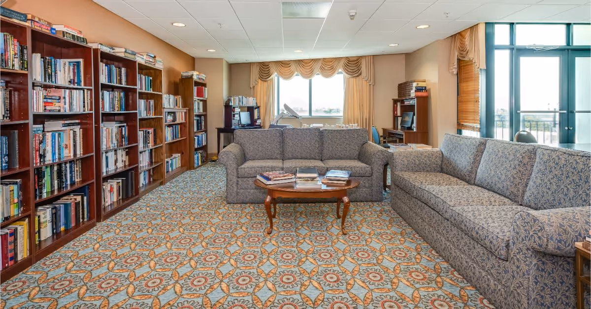 A cozy library or lounge area with patterned carpet, two large upholstered sofas facing each other with a wooden coffee table in between holding several books. Along the left wall are tall wooden bookshelves filled with books. At the back, there is a window with draped curtains letting in natural light, and a desk with a computer and chair. To the right, there is a glass door leading outside.