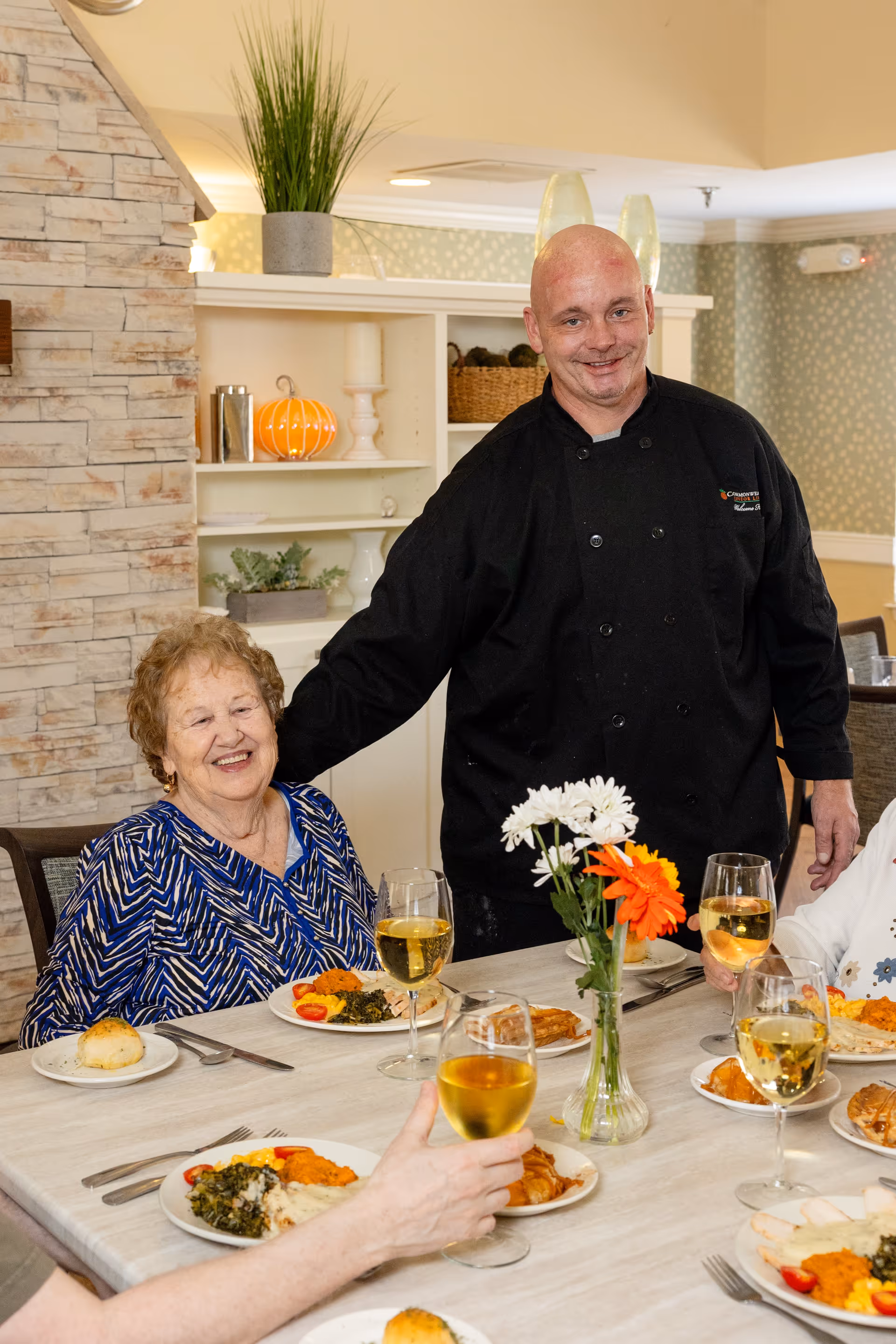 A smiling elderly woman seated at a dining table with plates of food and glasses of white wine, accompanied by a chef in a black uniform standing beside her in a warmly decorated dining room.