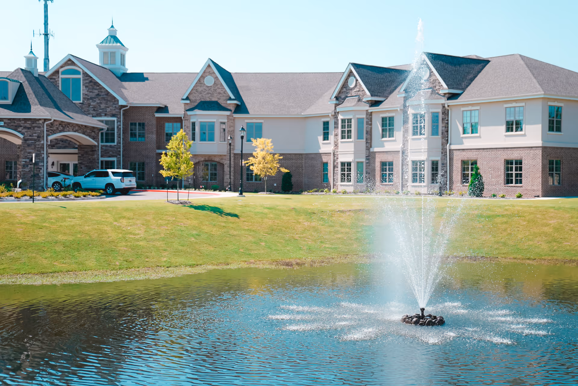 A large senior living facility building with a brick and stone exterior, multiple windows, and a peaked roof. In front of the building is a well-maintained grassy area with a pond featuring a water fountain spraying water upwards. There are a few small trees and parked cars near the entrance.