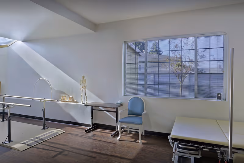 A bright rehabilitation room with parallel walking bars, a treatment table, a blue chair, and a large window.