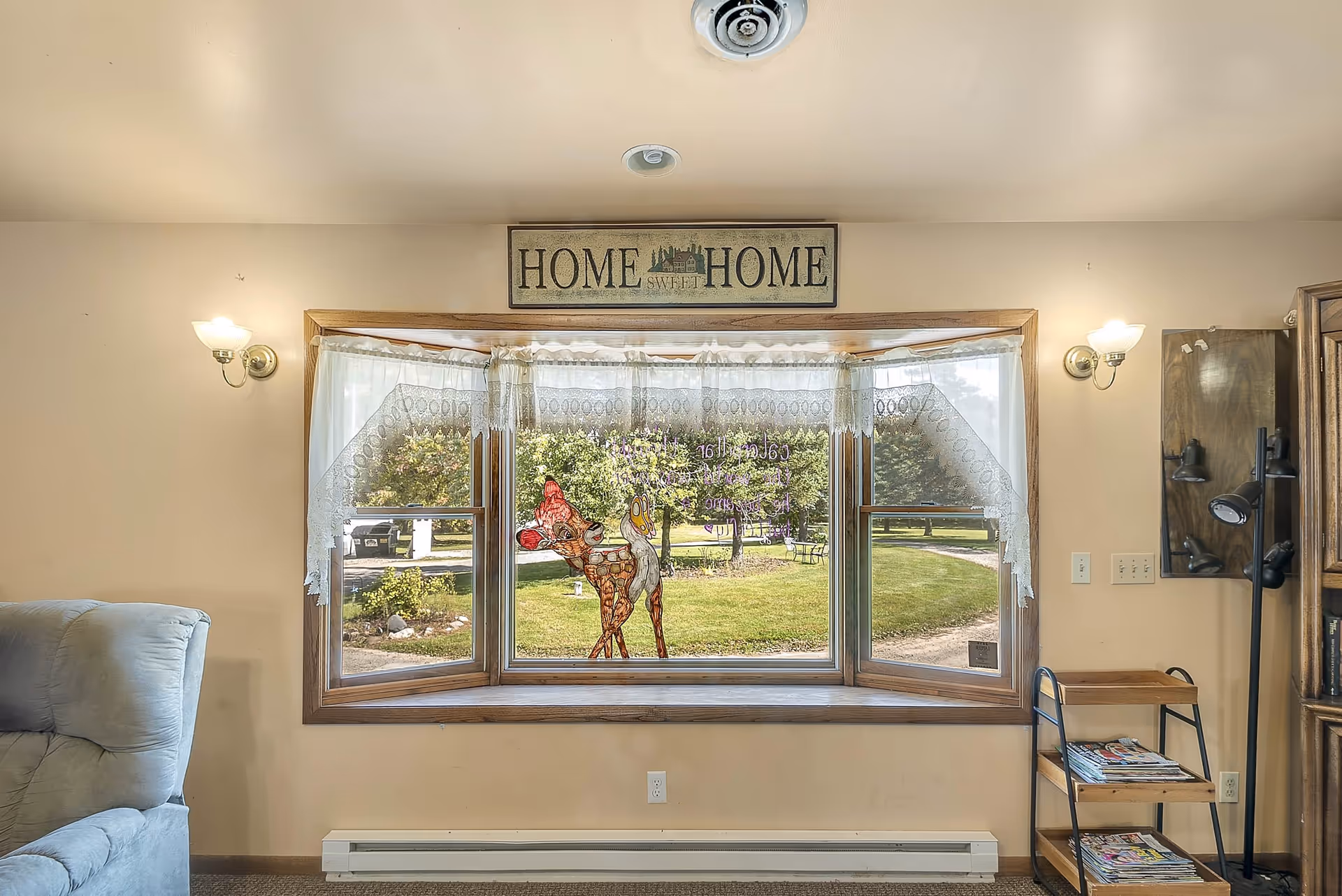 Interior view of a cozy living room area featuring a large bay window with lace curtains and a decorative stained glass deer. Above the window is a wooden sign that reads 'HOME SWEET HOME'. To the left is a light blue upholstered armchair, and to the right is a small wooden shelving unit with magazines and a black floor lamp. Outside the window, a green lawn with trees and a pathway is visible.