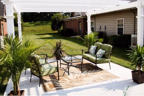 Outdoor patio with green cushioned chairs and a glass-top table under a white pergola overlooking a grassy yard.