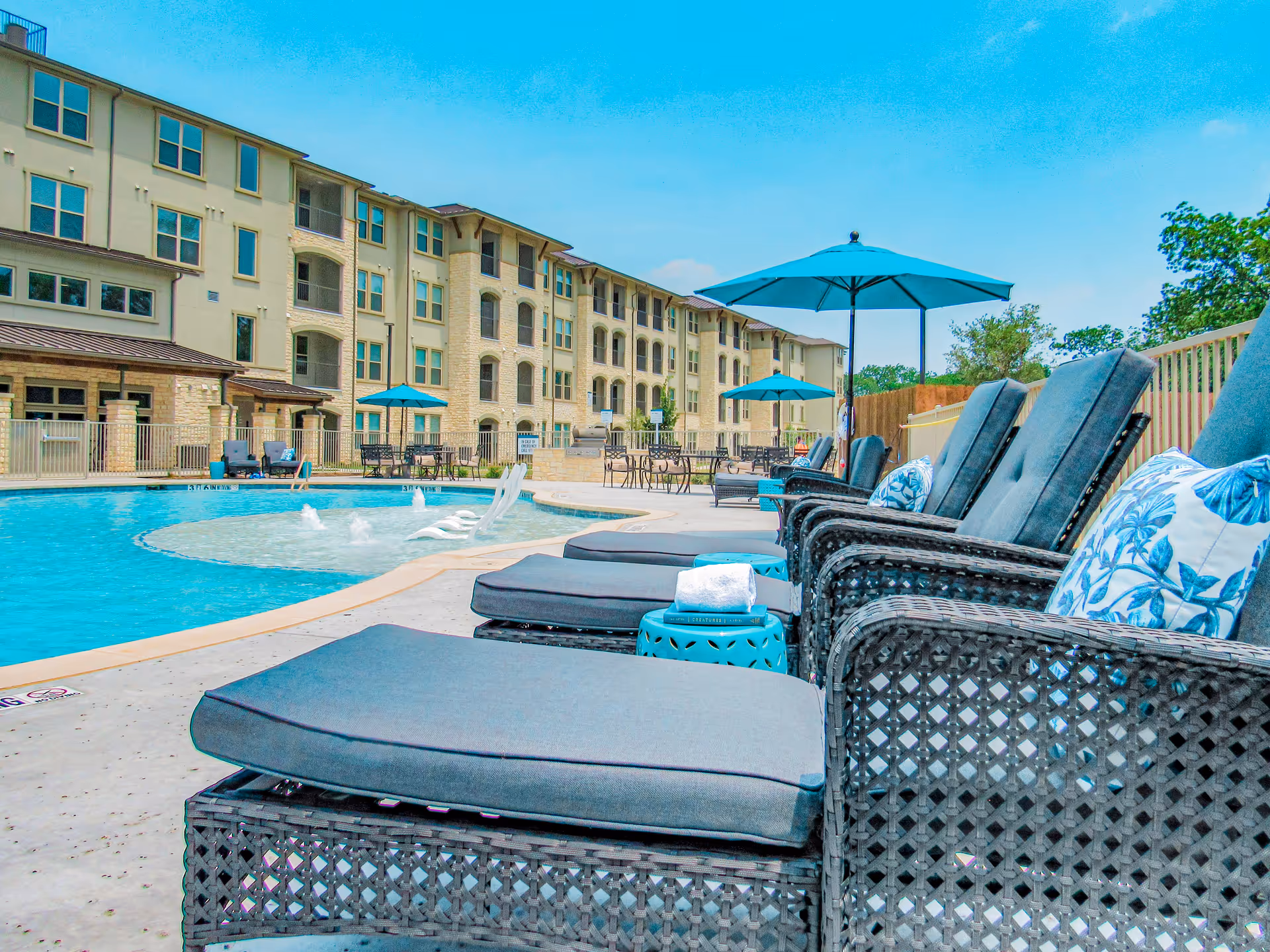 Outdoor pool area at Preserve at Willow Park with lounge chairs featuring blue cushions and floral pillows, blue umbrellas, and a multi-story residential building in the background under a clear blue sky.