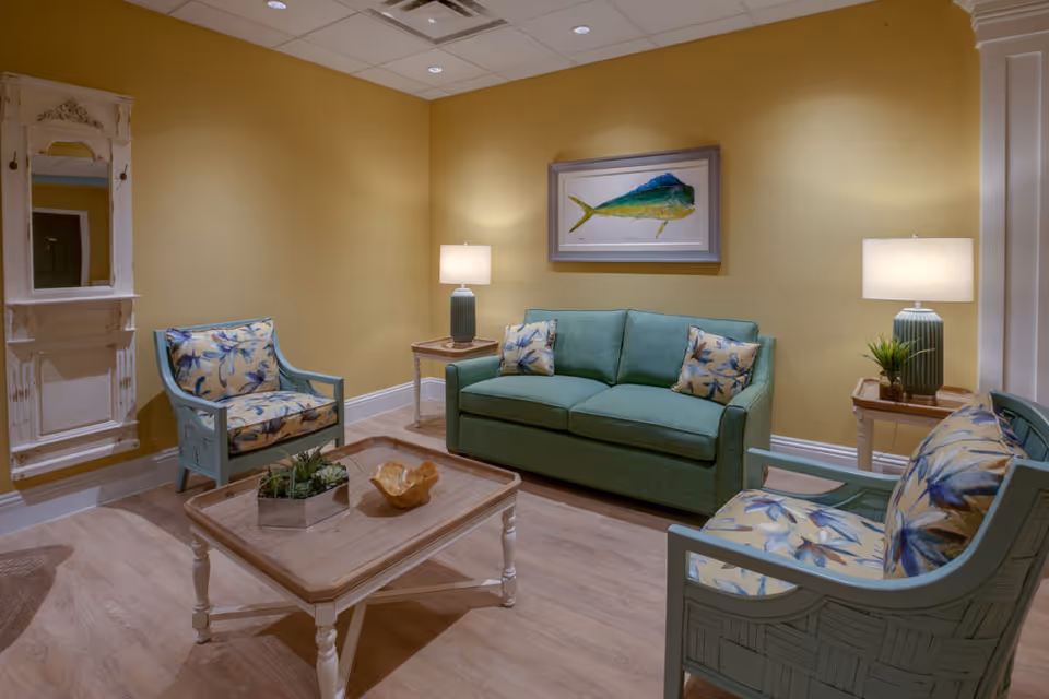 A cozy living room with yellow walls, featuring a green sofa with floral cushions, two matching armchairs with floral upholstery, a wooden coffee table with decorative items, two side tables each with a lamp and a small plant, and a framed painting of a fish on the wall.