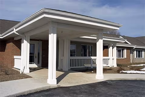 Exterior view of the entrance to a single-story brick building with white columns and a covered porch area. There is a paved driveway and some patches of snow on the ground.