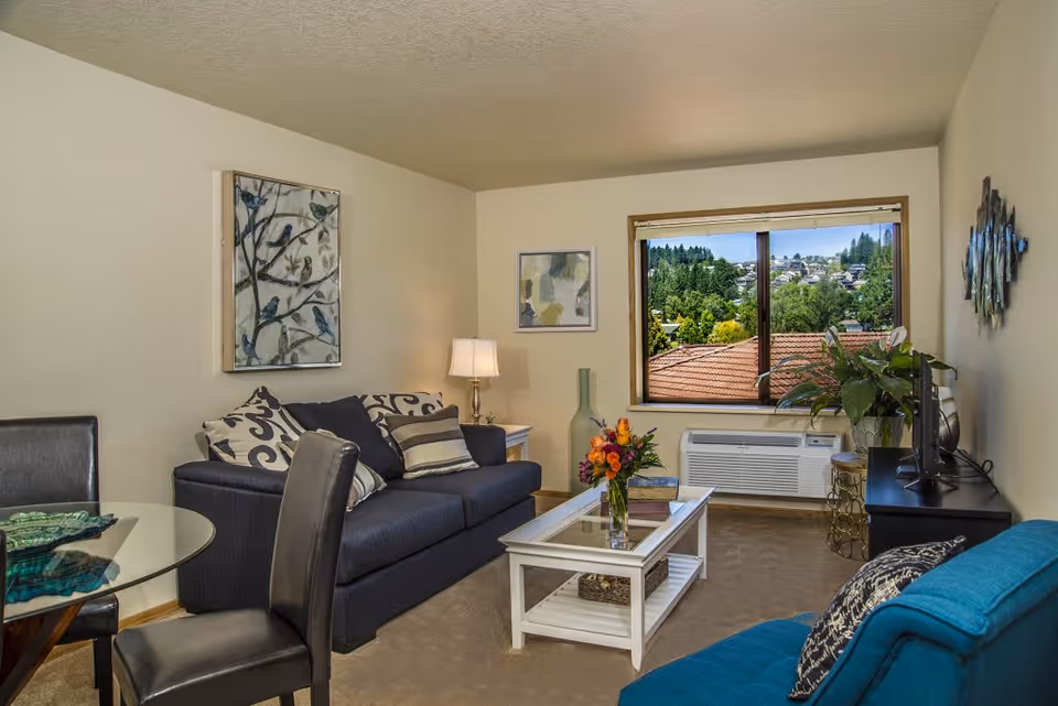A cozy living room in a senior living facility featuring a dark blue sofa with patterned cushions, a glass-top dining table with black chairs, a white coffee table with a vase of flowers, a blue armchair, a TV on a black stand, and a window showing a view of trees and rooftops outside.