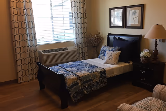 A furnished bedroom with a single dark wood bed, patterned bedding, nightstand with lamp and floral arrangement, and a window with patterned curtains.