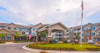 Exterior view of a large, multi-story senior living facility with beige and brown siding, multiple windows, and a covered entrance. There are two American flags on flagpoles in front of the building, a circular driveway, landscaped greenery, and a partly cloudy sky.