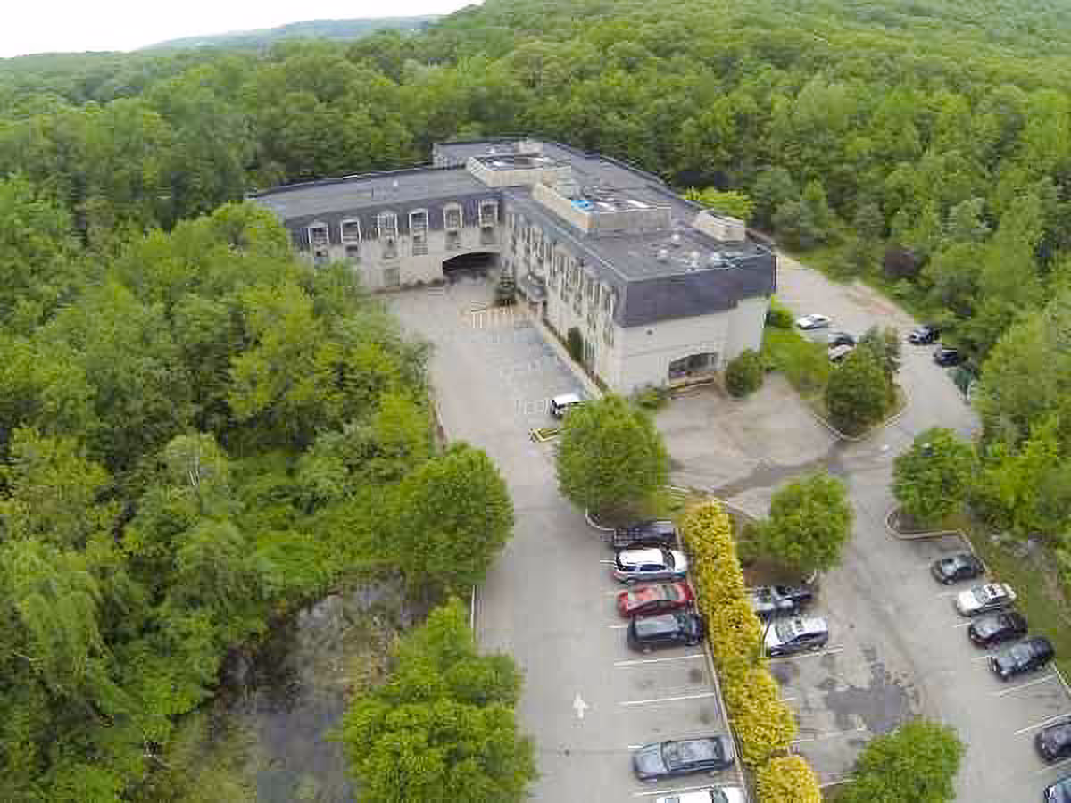 Aerial view of a large building surrounded by dense green trees and a parking lot with several cars parked. The building has a flat roof and multiple windows, situated in a forested area.