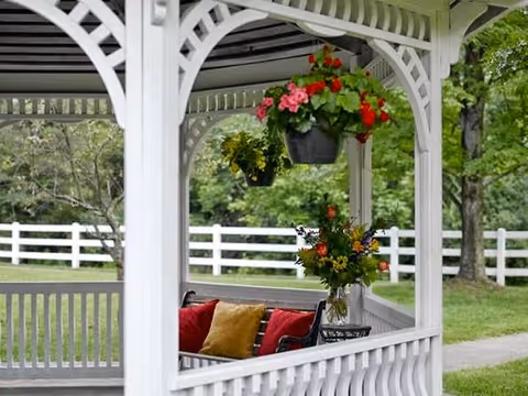 White wooden gazebo with hanging flower baskets and a bench with colorful pillows overlooking a fenced lawn.