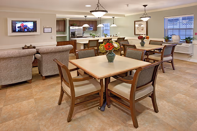 A well-lit common area in a senior living facility featuring several square tables with wooden chairs arranged around them. Each table has a vase with colorful flowers. In the background, there is a kitchen area with pendant lights, stainless steel appliances, and wooden cabinets. To the left, there are upholstered armchairs facing a wall-mounted television. Large windows with blinds allow natural light into the room.