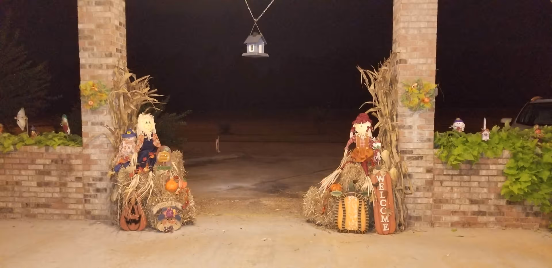 Nighttime view of a brick entrance with two pillars decorated with autumn-themed scarecrows, cornstalks, pumpkins, and a wooden welcome sign. A small birdhouse hangs from the top center between the pillars. Green plants are visible on the brick walls on either side.