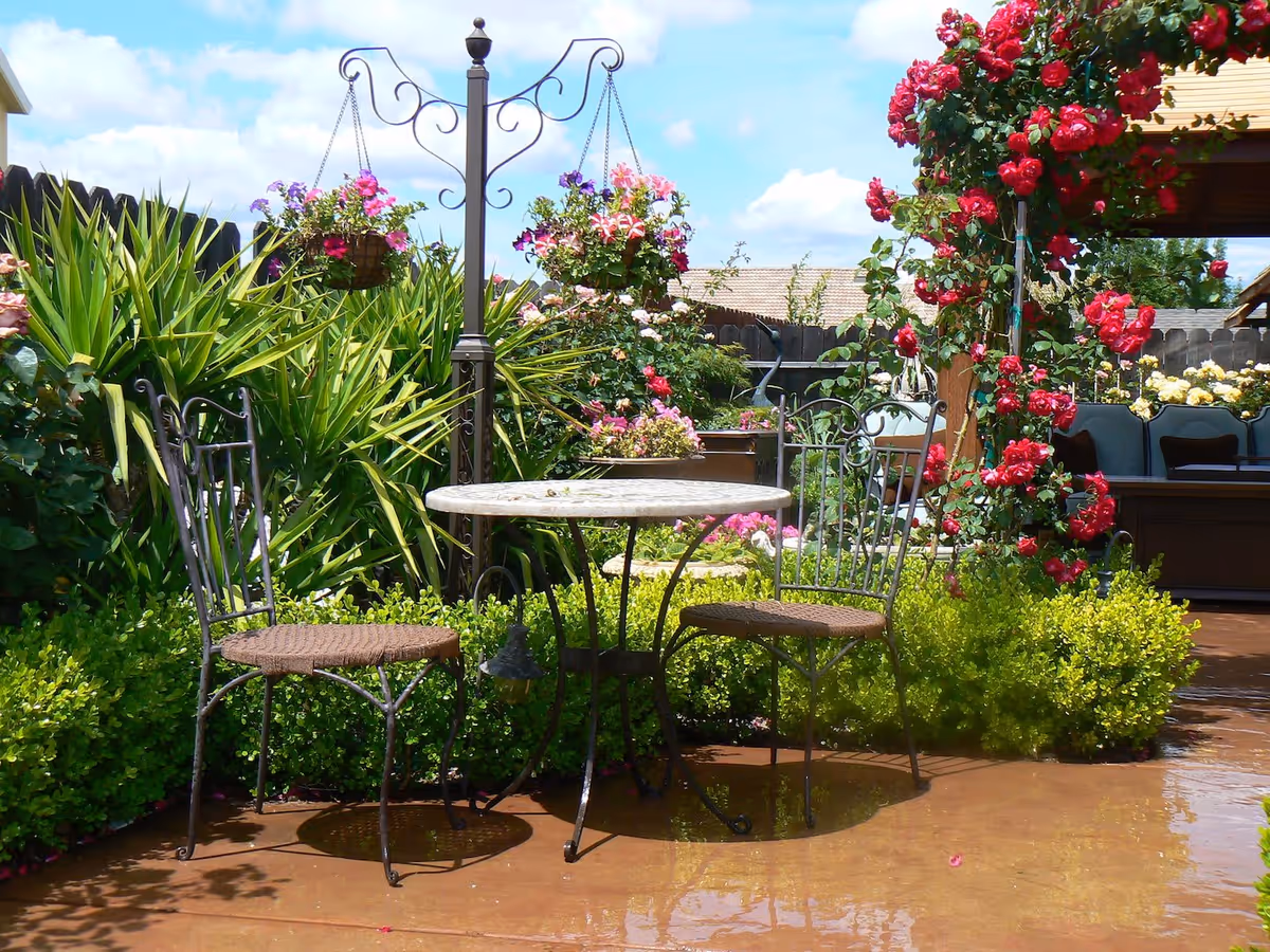A peaceful outdoor garden area with a small round table and two metal chairs with woven seats. The garden is lush with green plants and colorful flowers, including red roses climbing a wooden pergola. The sky is blue with some clouds, and the area appears sunny and inviting.